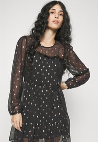 Μαύρο διάφανο φόρεμα με μακριά μανίκια, ριχτό ντεκολτέ και μεταλλική ροζ χρυσή σημειολογία. Διαθέτει ζώνη στη μέση και ριχτή εσοχή.