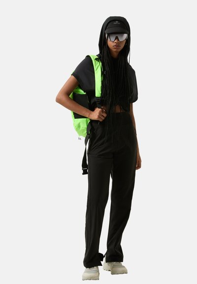 Schwarzes Cropped Top, schwarze weit geschnittene Hose, grüne Umhängetasche, graue Sneaker, schwarze Mütze und Sonnenbrille. Geflochtener Haarschnitt, minimalistisches Design.