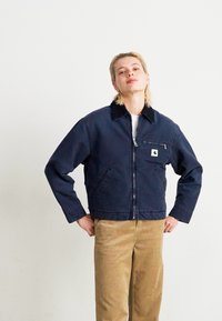Person with blonde hair wearing a navy blue zip-up jacket and beige corduroy pants, standing with hands on hips against a plain background.