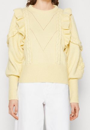 Lysegul strikket sweater med flæsedetaljer på skuldrene, flettemønstre foran og puffede ærmer, båret med hvide bukser.