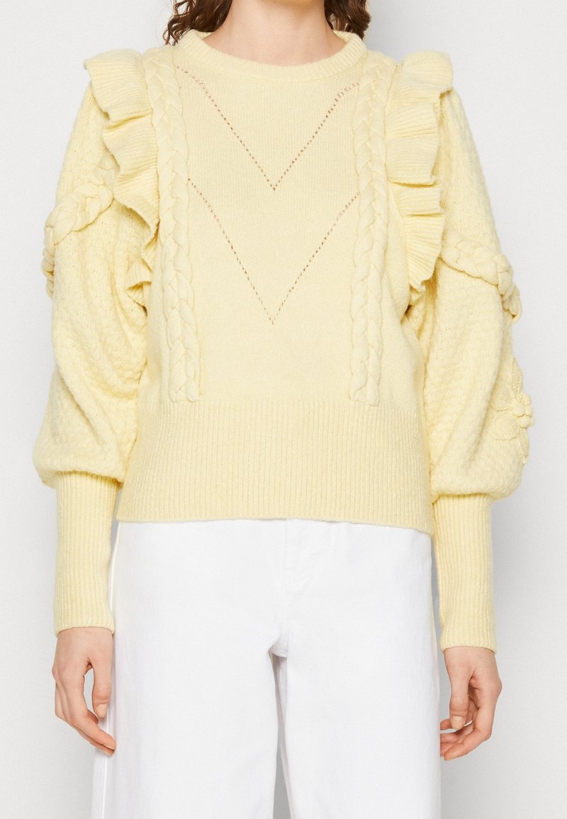 Pull en maille jaune clair avec des détails froncés aux épaules, des motifs tressés sur le devant et des manches bouffantes, porté avec un pantalon blanc.