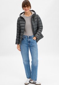 Vrouw die staat met één hand in de pocket, gekleed in een grijze pufferjack, lichtgrijs shirt, blauwe jeans, witte sneakers en een bril.