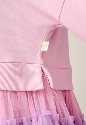 Roze top met lange mouwen en een geribbelde zoom, gedragen over een gelaagde roze tule rok, met een klein beige label aan de zijnnaad met de tekst "Little Bird".