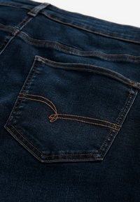 Zadné vrecko na tmavomodrých rifliach so žltým kontrastným prešitím a zaobleným dvojitým dekoratívnym dizajnom.