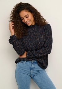 Tmavomodrá blúzka s paisley vzorom v svetlohnedej farbe; má vysoký golier a nariasené manžety; kombinovaná so svetlomodrými džínami.