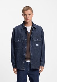 Camisa de mezclilla oscura con cierre de botones, dos bolsillos en el pecho y costuras contrastantes. Usada sobre un suéter marrón de punto y una camiseta blanca.