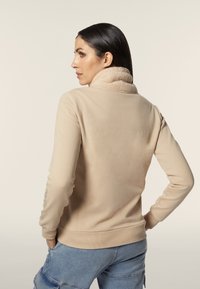 Giacca in pile beige con colletto alto, polsini e orlo a coste. Presenta una texture liscia e un sottile dettaglio di cucitura lungo le maniche.