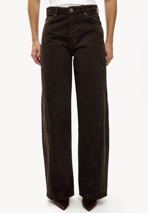 Femme portant un jean taille haute à jambes larges de couleur marron foncé avec des coutures avant, associé à des chaussures marron à bouts pointus et un haut blanc sans manches.