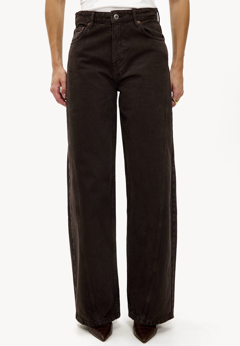 Femme portant un jean taille haute à jambes larges de couleur marron foncé avec des coutures avant, associé à des chaussures marron à bouts pointus et un haut blanc sans manches.