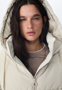 Doudoune beige avec une grande capuche, une fermeture éclair à l'avant et un cordon de serrage, portée sur un haut marron clair. Texture lisse et design minimaliste.