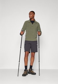 Oliven grøn fleece trøje med kvart lynlås, sort T-shirt under, grå shorts, sorte vandresko med orange detaljer, holder vandrestativer.