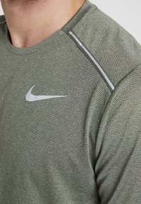 Zelené sportovní tričko vyrobené z texturované, prodyšné tkaniny. Obsahuje stříbrné logo Nike a reflexní prvky na švech ramen.