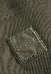Trest de culoare verde măsliniu cu un finisaj mat, având o pată pătrată marcată cu 'GABBA' în litere brodate și detalii de cusătură contrastante.