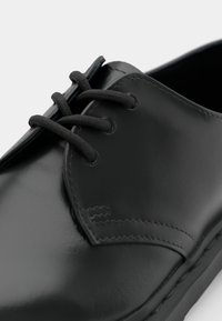 Musta nahkakenkä, jossa on sileä, kiiltävä pinta, pyöreä kärki ja näyttävät mustat nauhat pyöreissä silmukoissa. Hienovarainen ompelu yksityiskohtana.