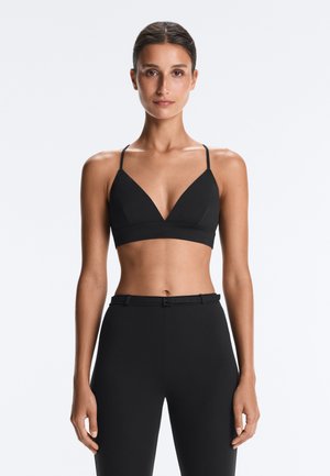 Femme debout face à l'avant portant un bralette noir avec des bretelles fines et un pantalon noir taille haute avec une ceinture.