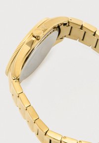 Gold metal wristwatch band and case, side view showing the crown and clasp on a light background.
