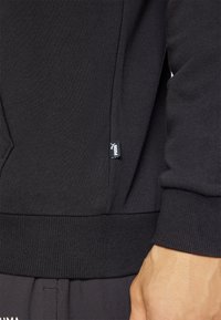 Black sweatshirt with ribbed cuffs and hem, featuring a small white logo tag on the side. Soft fabric with a smooth texture.