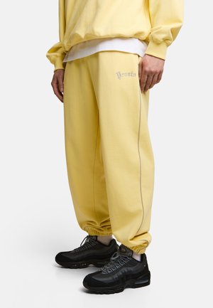 Osoba ubrana w żółte spodnie dresowe z napisem "Prosto", pasującą żółtą bluzę, białe skarpetki oraz czarne buty Nike Air Max.