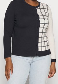Woman wearing a buttoned cardigan with a black solid left side and white right side featuring a black grid pattern, paired with light blue jeans.