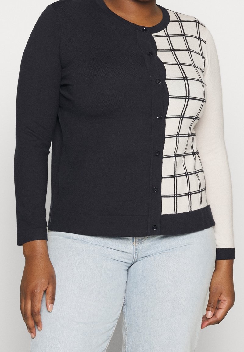 Woman wearing a buttoned cardigan with a black solid left side and white right side featuring a black grid pattern, paired with light blue jeans.