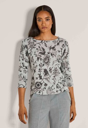 Vrouw met lang, donker haar in een grijs bloemdessin langemouwen top en lichtgrijze broek, poserend tegen een effen lichte achtergrond.