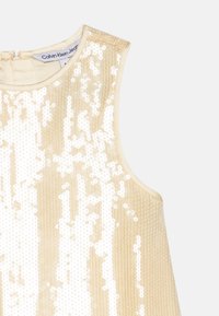 Haut sans manches beige présentant un motif de sequins avec des rayures verticales, un col rond et une fermeture dans le dos. Étiquette de marque visible.
