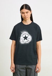 Juodas medvilninis marškinėliai su centralia balta grafika, kuriame pavaizduota žvaigždė ir užrašai "CONVERSE" ir "ALL STAR". Trumpomis rankovėmis ir apvaliu kaklu.