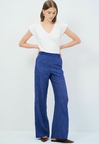 Pantalones de pierna ancha de encaje azul con un patrón texturizado, combinados con una blusa blanca de escote en V lisa. Presentan una cinturilla azul y zapatos planos.