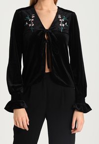 Svart sammetstopp med V-ringning, framslutning med knytning, långa ärmar med utsvängda manschetter och blommönster i rosa och grönt.