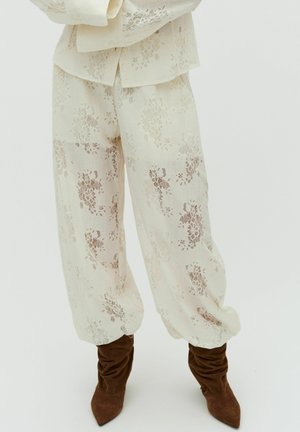 Loose cream lace pants with floral patterns worn with brown slouchy pointed-toe boots on a plain background.