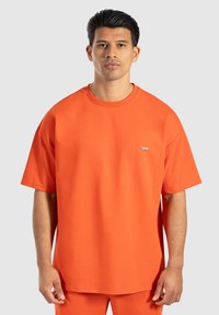 Oranžové oversized tričko z měkké bavlny, s kulatým výstřihem a malým logem na hrudi. Hladká textura, krátké rukávy.