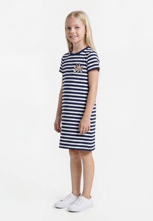 Jeune fille debout et souriante, portant une robe à manches courtes rayée bleu marine et blanche avec un motif de soleil et des baskets blanches.