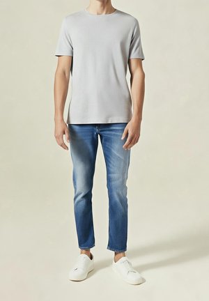 Homme portant un t-shirt à manches courtes gris clair, un jean slim bleu délavé et des baskets blanches, debout devant un fond uni.