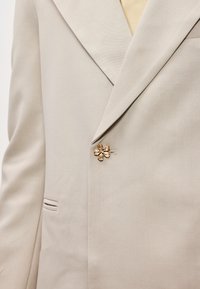 Blazer bege com lapela acentuada, tecido texturizado e um broche em forma de flor dourada no lado esquerdo. Apresenta um bolso lateral.