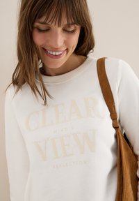 Sweatshirt blanc avec un texte en relief beige : "REFLET DE L'ESPRIT CLAIR." Matière douce, décolleté rond, associé à une sangle de sac en suède marron.