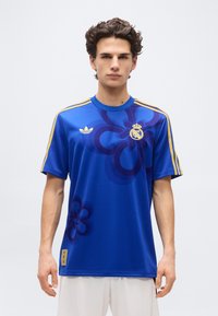Young man wearing a royal blue sports jersey with gold stripes on sleeves and Real Madrid and Adidas logos, standing against plain white background.