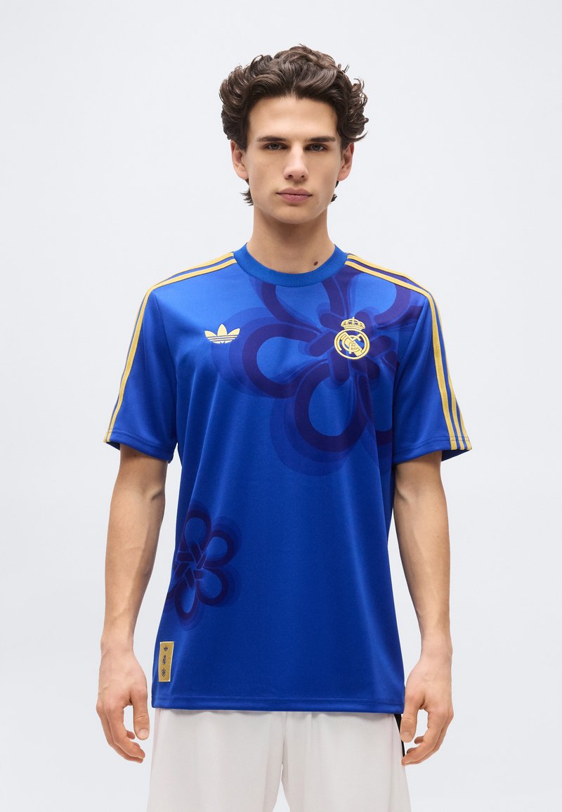 Young man wearing a royal blue sports jersey with gold stripes on sleeves and Real Madrid and Adidas logos, standing against plain white background.