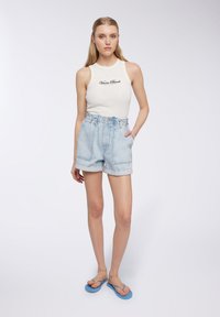 Top bianco senza maniche con la scritta "Venice Beach" stampata in nero, abbinato a pantaloni corti di denim azzurri a vita alta e ciabatte blu.