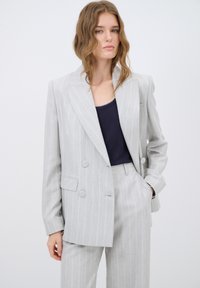 Lichtgrijs streepjespak met een dubbelgeknoopte blazer met grote knopen en bijpassende broek. De basislaag is een donkerblauwe top.