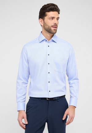 Helles blaues Langarmhemd, strukturierter Stoff, Hemdkragen zum Knöpfen, mit dunklen Knöpfen und einer Brusttasche, kombiniert mit dunklen Hosen.