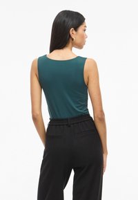Top senza maniche color verde petrolio con scollo rotondo, texture liscia, abbinato a pantaloni neri con vita elastica e tasche laterali.