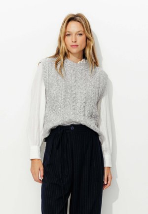 Un gilet en tricot gris avec un motif en câble sur une chemise à manches longues blanche. Les pantalons sont sombres avec des rayures verticales, dotés d'une taille nouée.