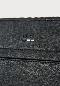 Czarny portfel z fakturowanej skóry z zamkiem błyskawicznym. Posiada wytłoczone logo "BOSS" oraz trzy małe metalowe akcenty w kolorze srebrnym, białym i brązowym.