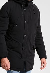 Manteau rembourré noir avec capuche, doté de boutons à l'avant, de deux poches latérales et d'une texture lisse. Accentué par des cordons de serrage à la capuche.