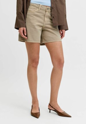 Femeie purtând pantaloni scurți bej din denim, sacou maro cu carouri, top bleu deschis și pantofi cu toc slingback maro, cu vârf ascuțit, pe un fundal neutru.