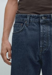 Jeans in denim blu scuro con taglio dritto, dotati di chiusura a bottone, passanti per cintura e dettagli di cucitura visibili. Mano appoggiata sulla vita.