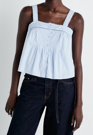 Dark-skinned person wearing a light blue sleeveless button-front top with lace trim and dark high-waisted jeans against a plain background.