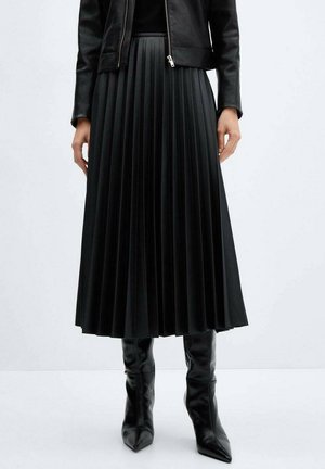 Femme portant une jupe midi plissée noire, une veste en cuir noire zippée et des bottes noires hautes jusqu’aux genoux à bout pointu sur fond uni.