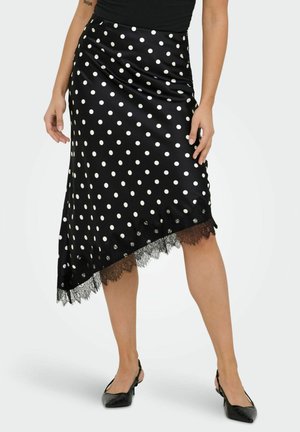 Kobieta ubrana w czarną asymetryczną spódnicę w groszki z koronkową lamówką oraz czarne, niskie buty na obcasie, stojąca na jednolitym tle.