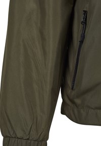 Gros plan sur la manche et le côté d'une veste vert olive avec un poignet élastique et une poche zippée noire visible.